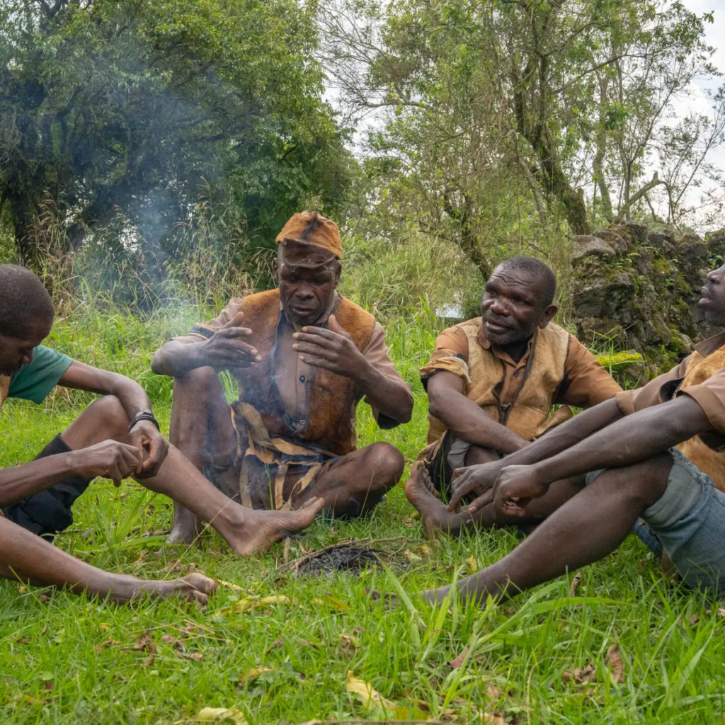 Four men in traditional-style clothing sitting together on grass near a small fire